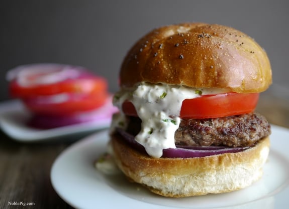 The Juciest Mediterranean Style Burgers with Creamy Feta Curry Dressing on a white plate with tomato and onion in the background.
