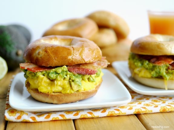 Two cheesy Egg Avocado and Bacon Breakfast Sandwich sitting on a white plates with bagels and juice in the background.