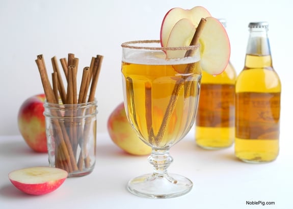 Beer Cider Slam in a tall glass with a cinnamon stick and a slice of apple. Cinnamon sticks in a jar, sliced and whole apples and two bottles of cider.