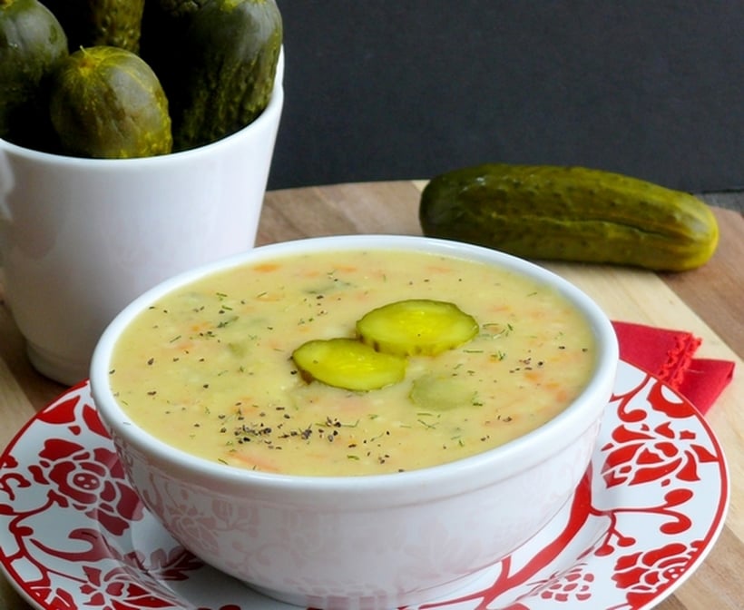 Bowl of dill pickle soup in a white bowl, sitting on a red and white plate with more pickles in the background.