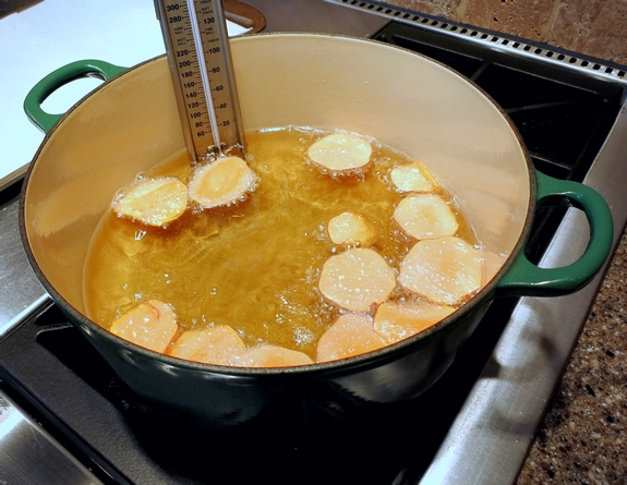Several sweet potato slices frying in hot oil with a candy thermometer.