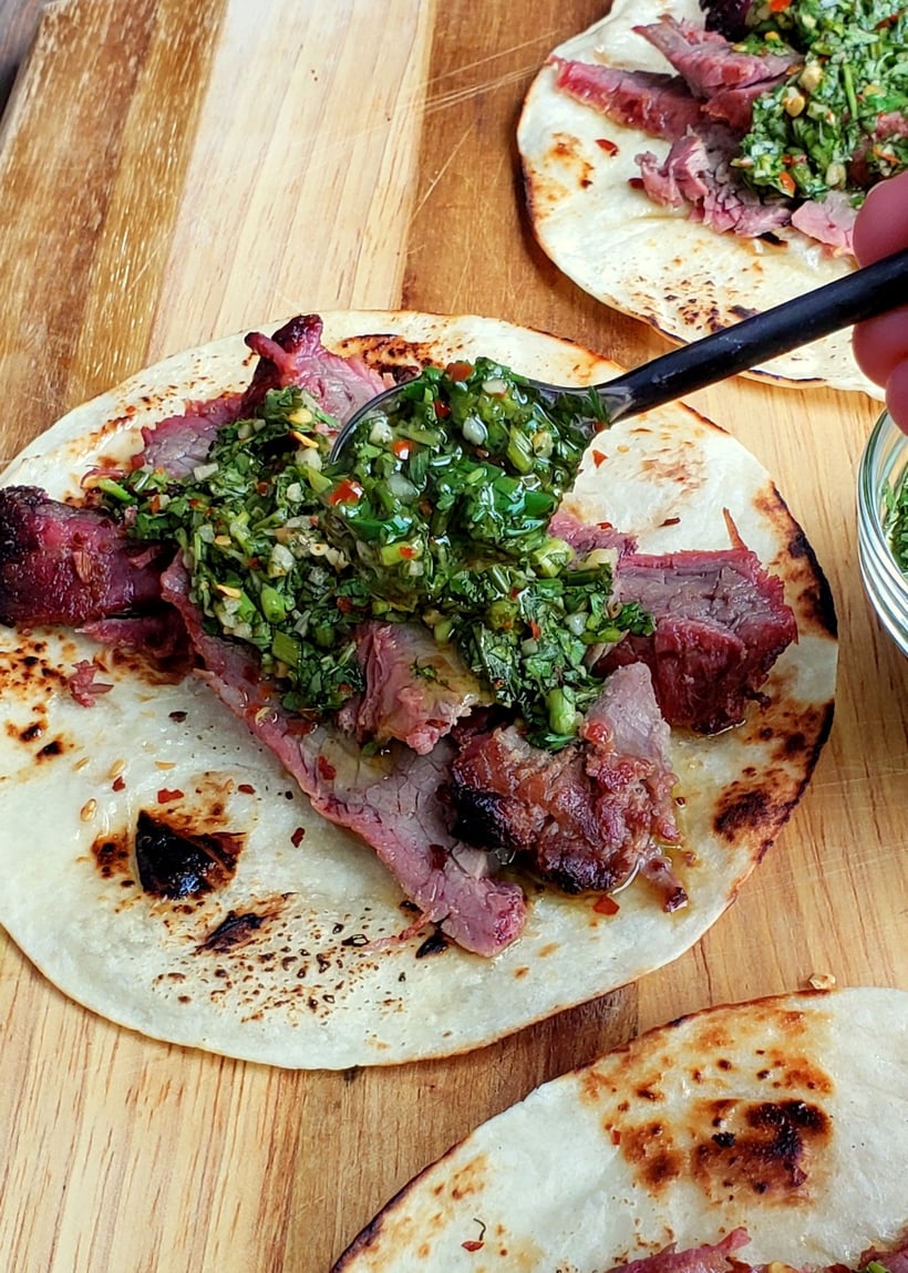 Chimichurri sauce being spooned on flank steak sitting on a tortilla.