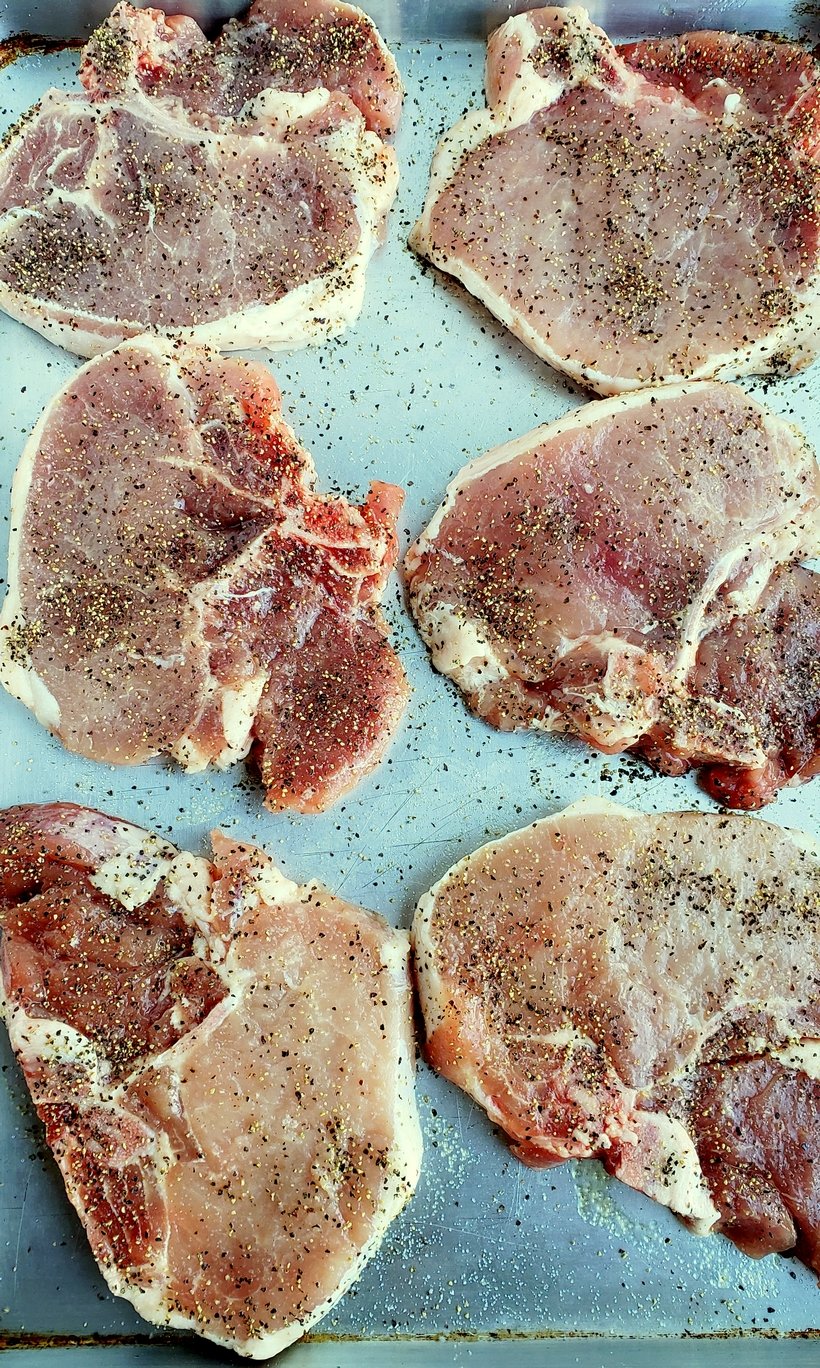 Six raw pork chops on a baking sheet seasoned with salt and pepper.