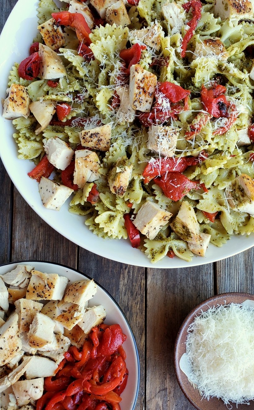 Chicken pesto pasta in a white bowl surrounded by a bowl of chicken and red peppers and grated parmesan cheese.