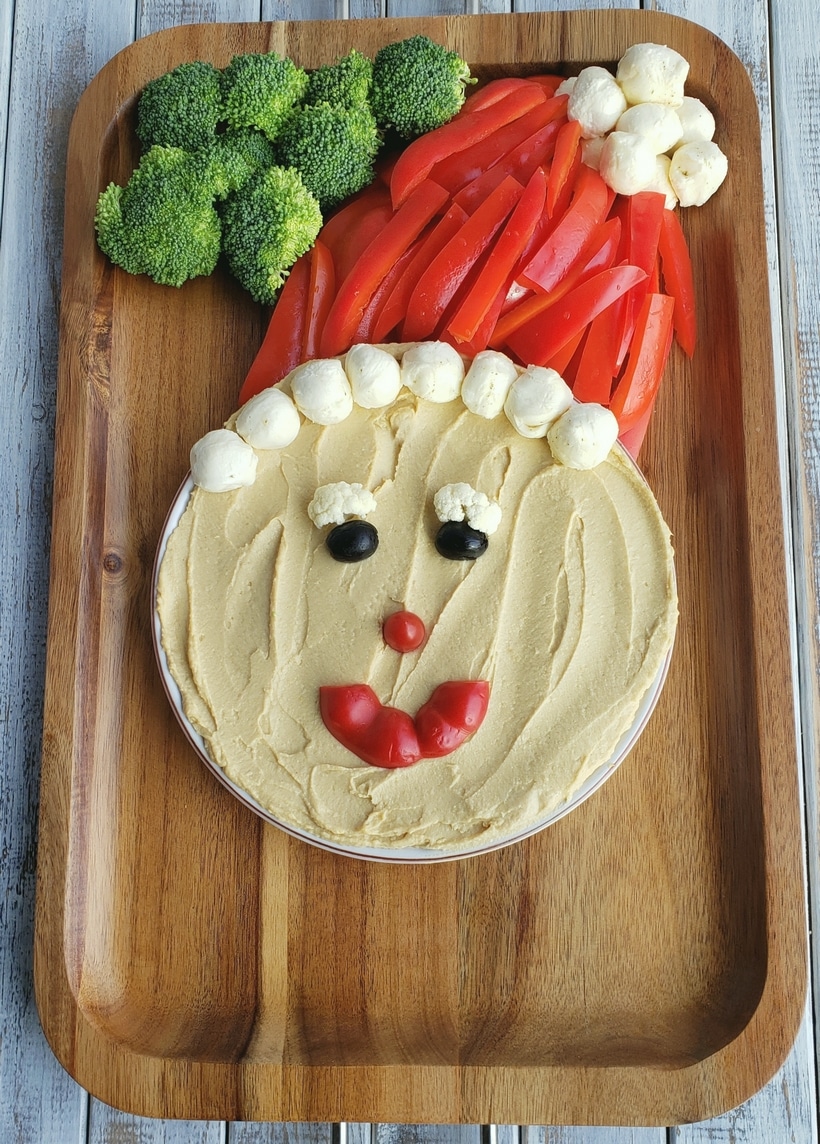Snack board being built to look like santa with hummus face, broccoli, red peppers and cheese.