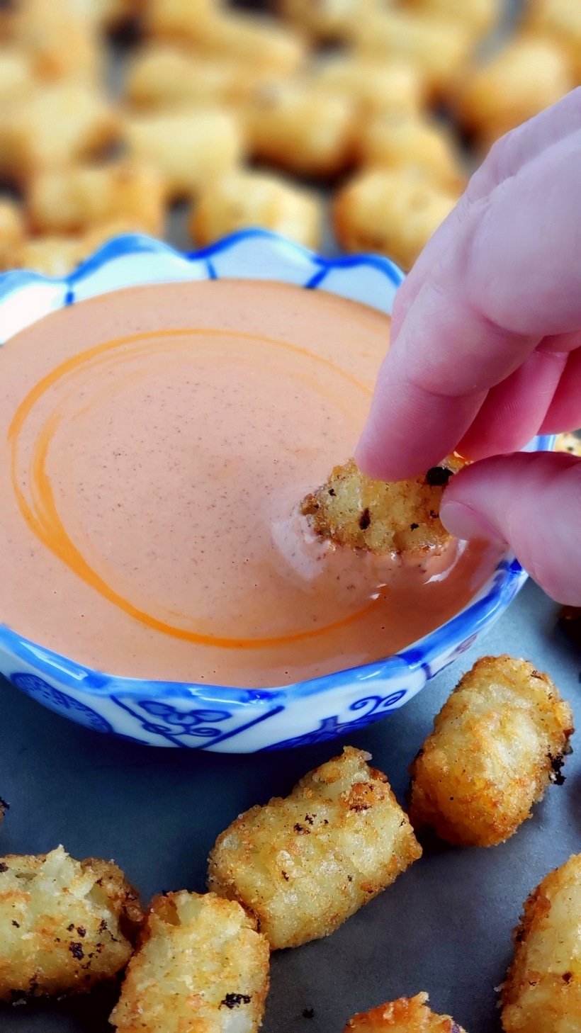 Hand dipping a tater tot into dipping sauce surrounded by tots.
