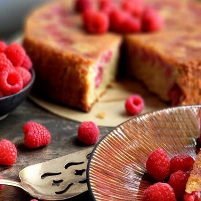 Cake with a slice missing surrounded by raspberries.