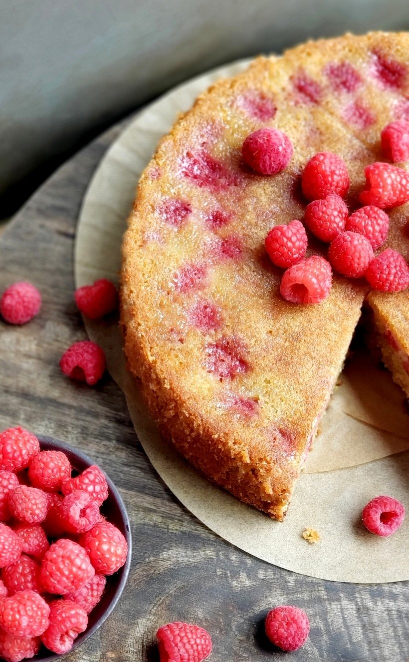 Whole raspberry cake with a slice missing. Fresh raspberries on the side and on top of the cake.