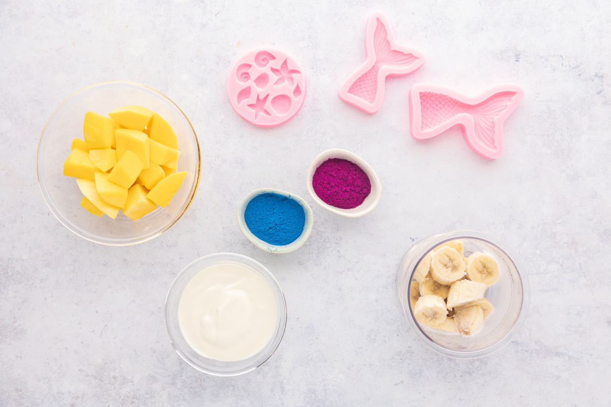Ingredients for mermaid smoothie bowls including frozen bananas, mango, yogurt, blue spirulina powder, beet root powder, and silicone mermaid molds.