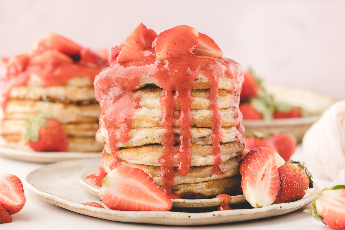 Strawberry buttermilk pancakes stacked on a plate with warm homemade strawberry sauce dripping down the sides and fresh strawberries around the stack.