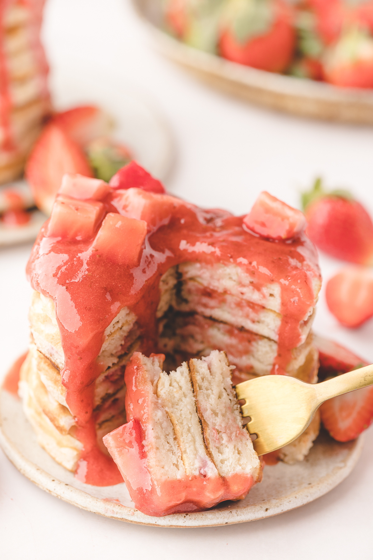 Fork lifting a bite of strawberry pancakes with jammy strawberries inside the buttermilk stack and warm strawberry sauce dripping over the top.