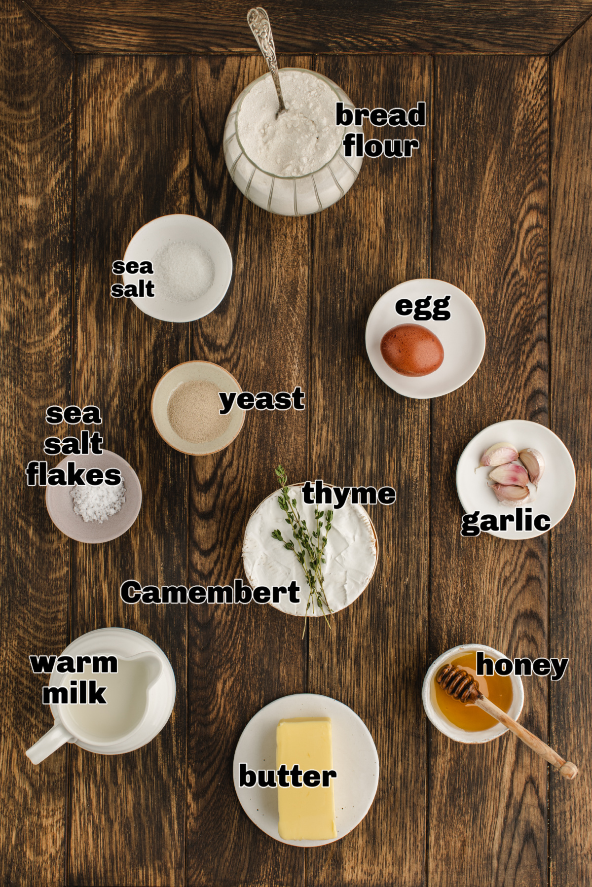 Ingredients for a baked Camembert bread roll wreath arranged on a wooden board, including flour, yeast, warm milk, butter, garlic, thyme, honey, egg, and sea salt.