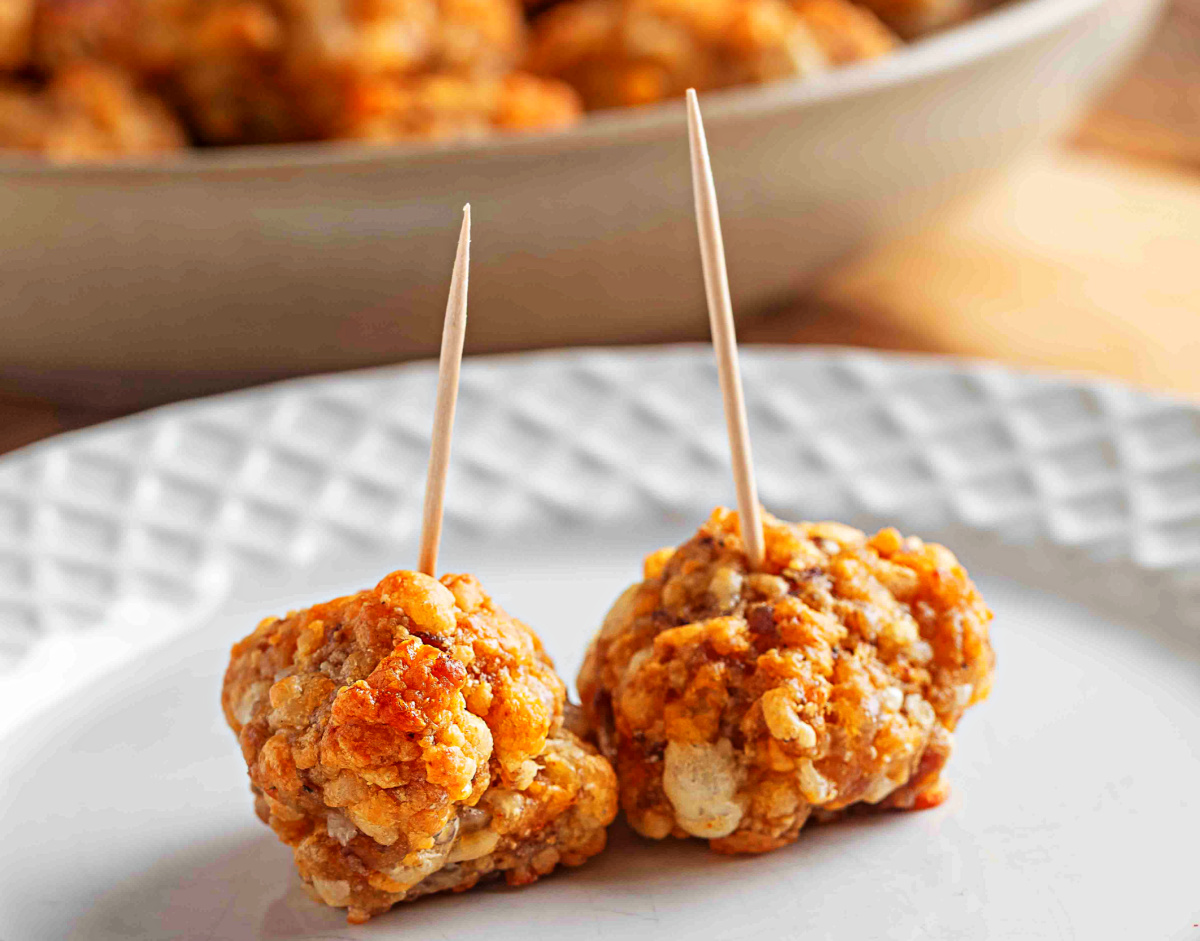 Two smoked cheddar sausage balls with toothpicks stand on a white plate, showing their crisp baked edges and tender Bisquick binding.