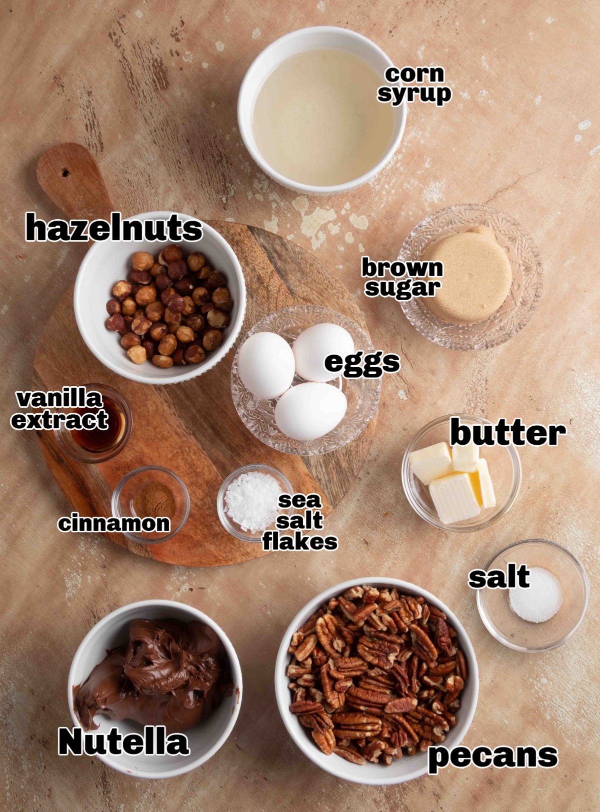 Ingredients for chocolate hazelnut pecan pie laid out on a board, including Nutella, pecans, hazelnuts, eggs, butter, brown sugar, corn syrup, cinnamon, vanilla, and sea salt.