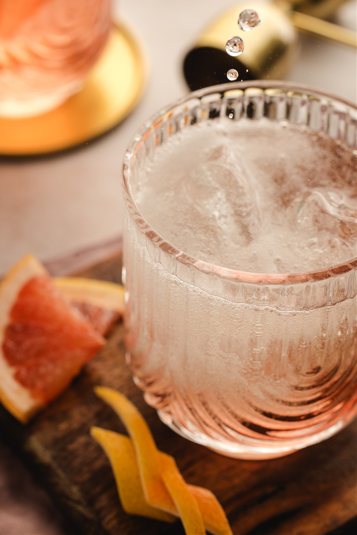 A chilled Lillet Ros&eacute; spritz catching falling droplets, the pale pink drink bubbling over ice with grapefruit slices and twists scattered on the wooden board below.