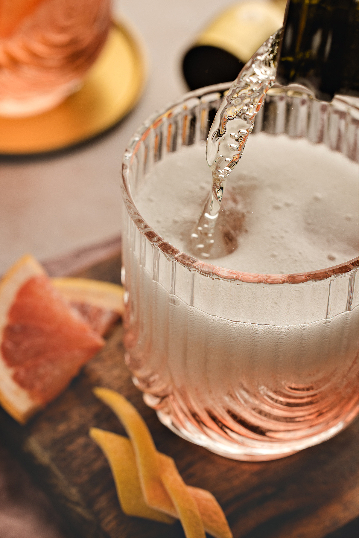 Sparkling wine being poured into a glass of Lillet Ros&eacute; spritz, creating a foamy layer over the pale pink drink with grapefruit slices and twists nearby.