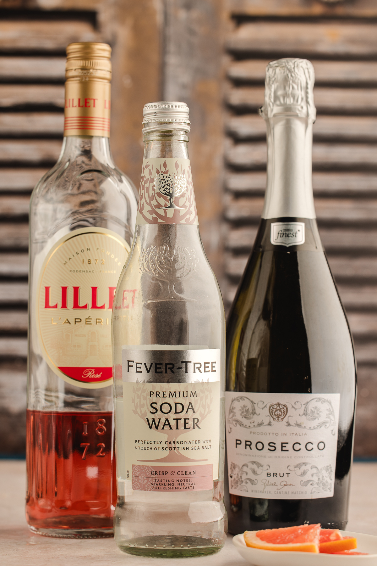 The three ingredients for a Lillet Ros&eacute; spritz&mdash;Lillet Ros&eacute;, club soda, and a bottle of brut prosecco&mdash;standing together with fresh grapefruit slices nearby.
