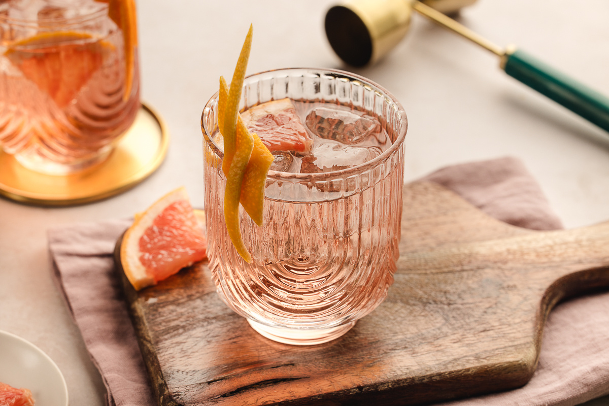 A pale pink Lillet Ros&eacute; spritz served over ice in a textured glass, garnished with a long grapefruit twist and set on a wooden board with fresh grapefruit nearby. 