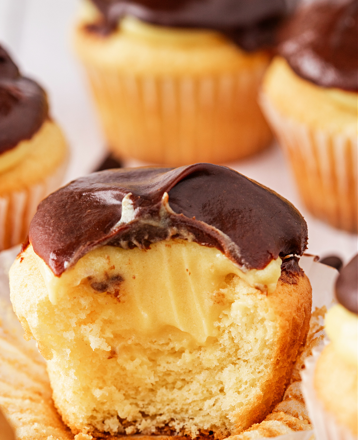 Close-up of a filled cupcake showing creamy vanilla pudding inside and a thick layer of chocolate ganache on top, with more cupcakes in the background