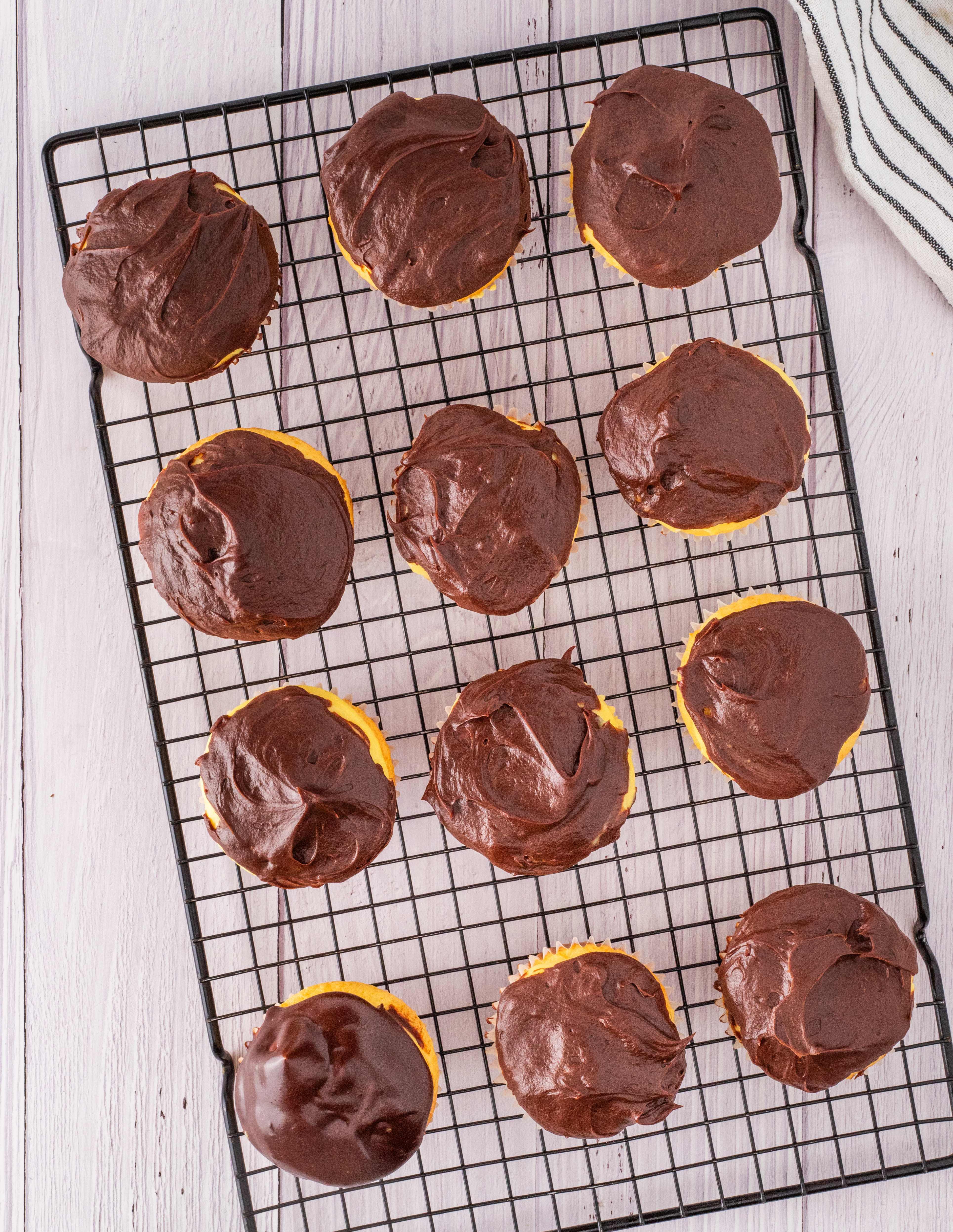 Freshly frosted cupcakes with chocolate ganache cooling on a wire rack, showing the thick glossy layer set over vanilla cream centers