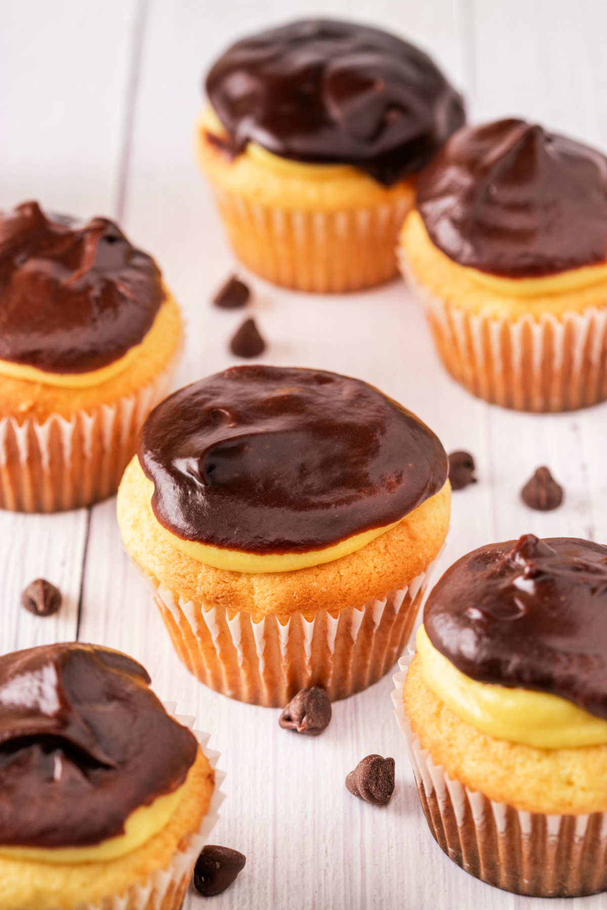 Group of cupcakes with vanilla pudding filling and smooth chocolate ganache topping, arranged on a white wood surface with chocolate chips scattered around
