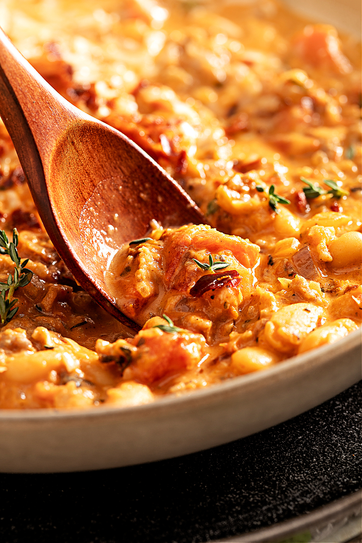 Tuscan-style white bean and sausage soup simmering with prosciutto, tomatoes, and thyme, stirred with a wooden spoon in a skillet.