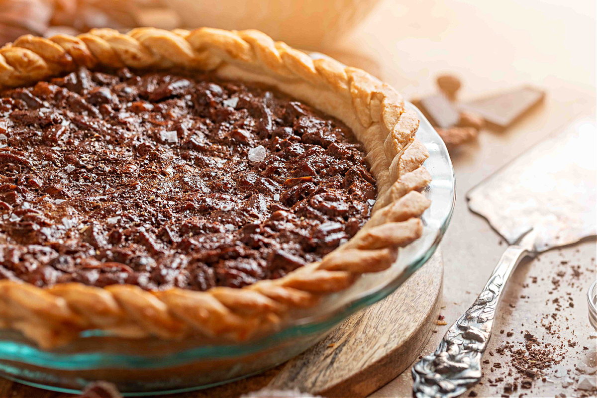 A full view of the baked chocolate hazelnut pecan pie showing the dark, molten filling and braided pie crust on a wooden board.