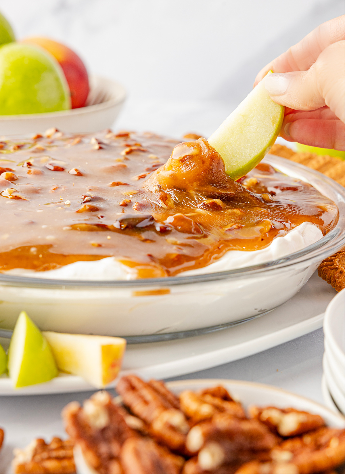 Pecan pie dip with a cold cream cheese base topped with warm caramel pecans as a green apple slice is dipped into the topping, showing the gooey texture by Noble Pig.