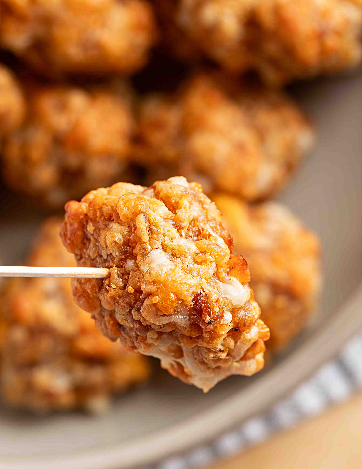 A single sausage ball held on a toothpick, showing its crisp, golden exterior and melted smoked cheddar against a blurred bowl of more sausage balls.