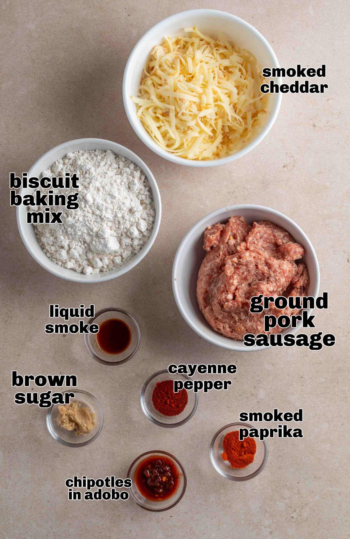 Ingredients for smoked cheddar sausage balls arranged on a countertop, including biscuit baking mix, ground pork sausage, shredded smoked cheddar, chipotle in adobo, brown sugar, smoked paprika, cayenne, and liquid smoke.