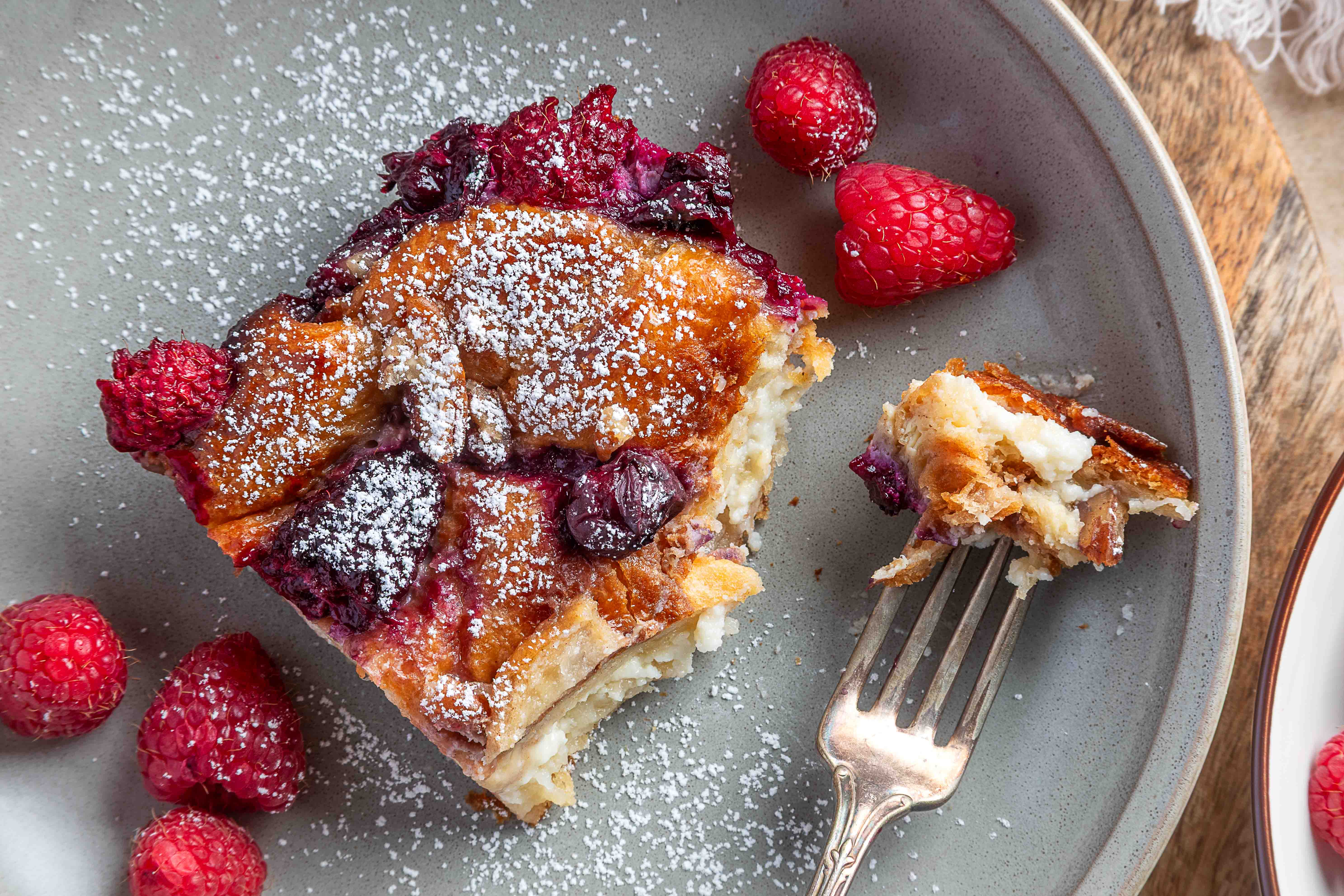 A spoon scoops into the baked croissant French toast casserole in the dish, showing the custardy interior with berries and pecans on top.