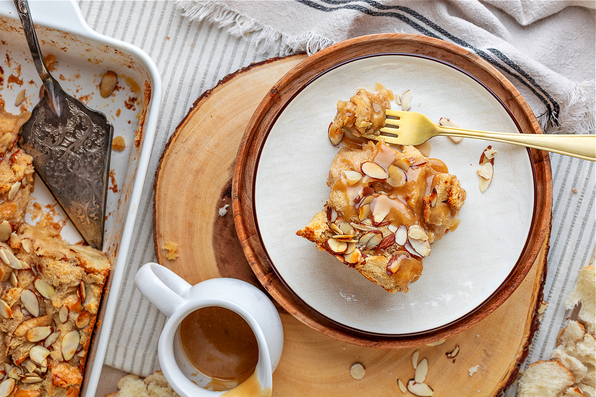 A slice of almond cream bread pudding on a plate with warm glaze and sliced almonds, shown overhead with a fork and the remaining pudding in the pan beside it.
