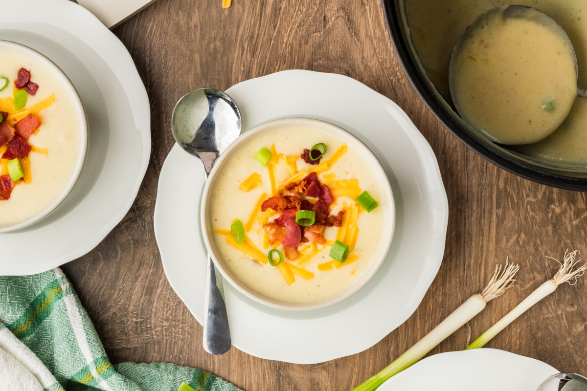 Bowls of creamy potato soup topped with crisp bacon, shredded cheddar, and fresh scallions on a wooden table.