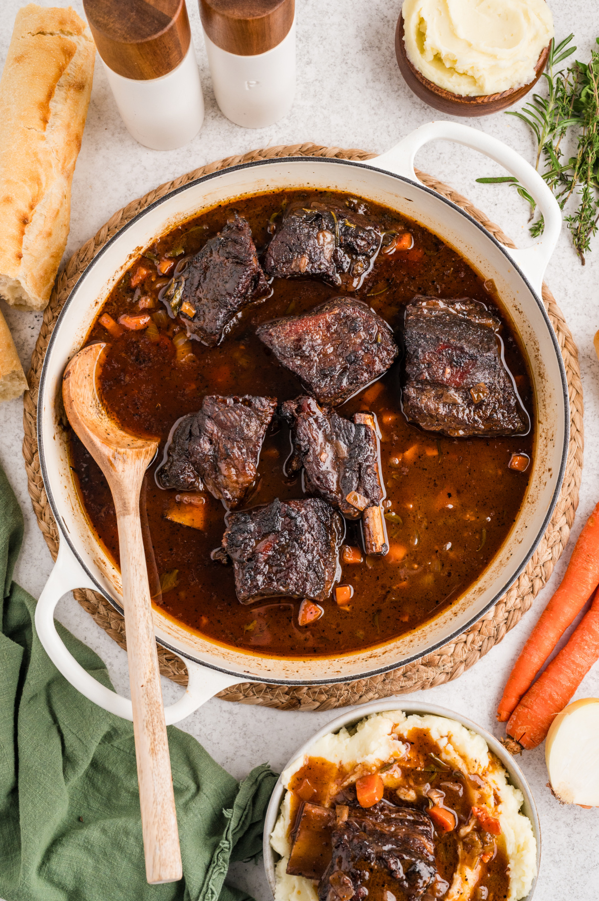 Short ribs braising in a wide pan with a dark, glossy sauce, vegetables, and herbs after hours in the oven.