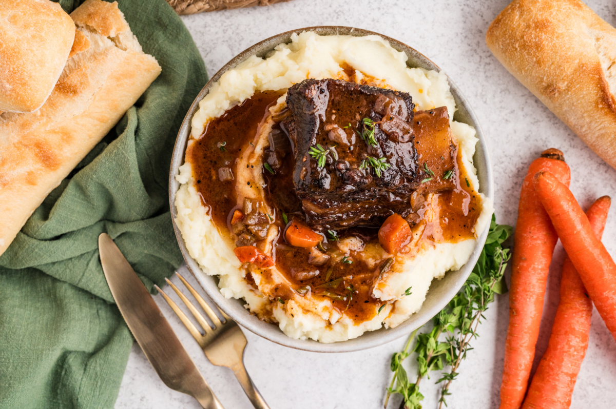 Braised beef short ribs served in a bowl of mashed potatoes with carrots, herbs, and a deep, savory sauce.
