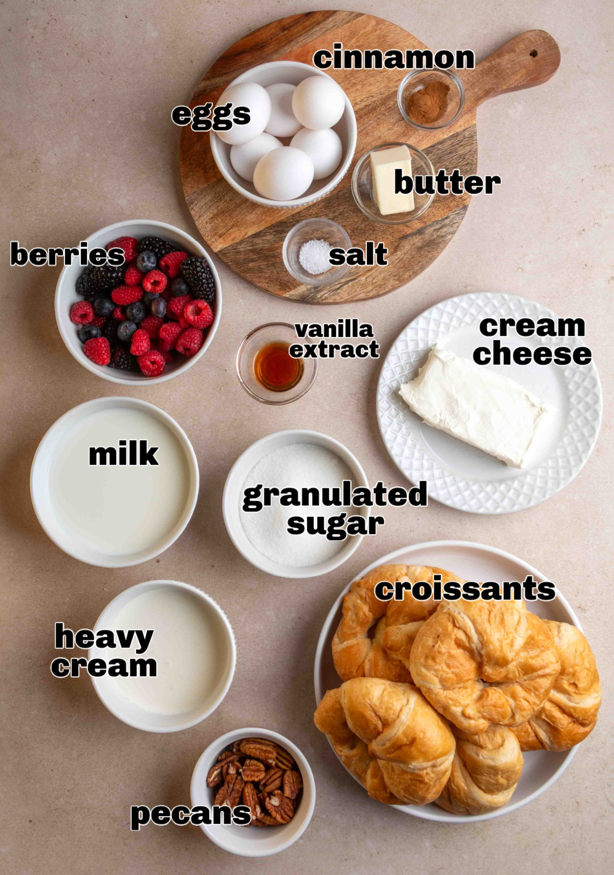 Ingredients for a croissant French toast casserole arranged on a counter, including croissants, cream cheese, eggs, berries, pecans, milk, heavy cream, sugar, butter, cinnamon, vanilla, and salt.