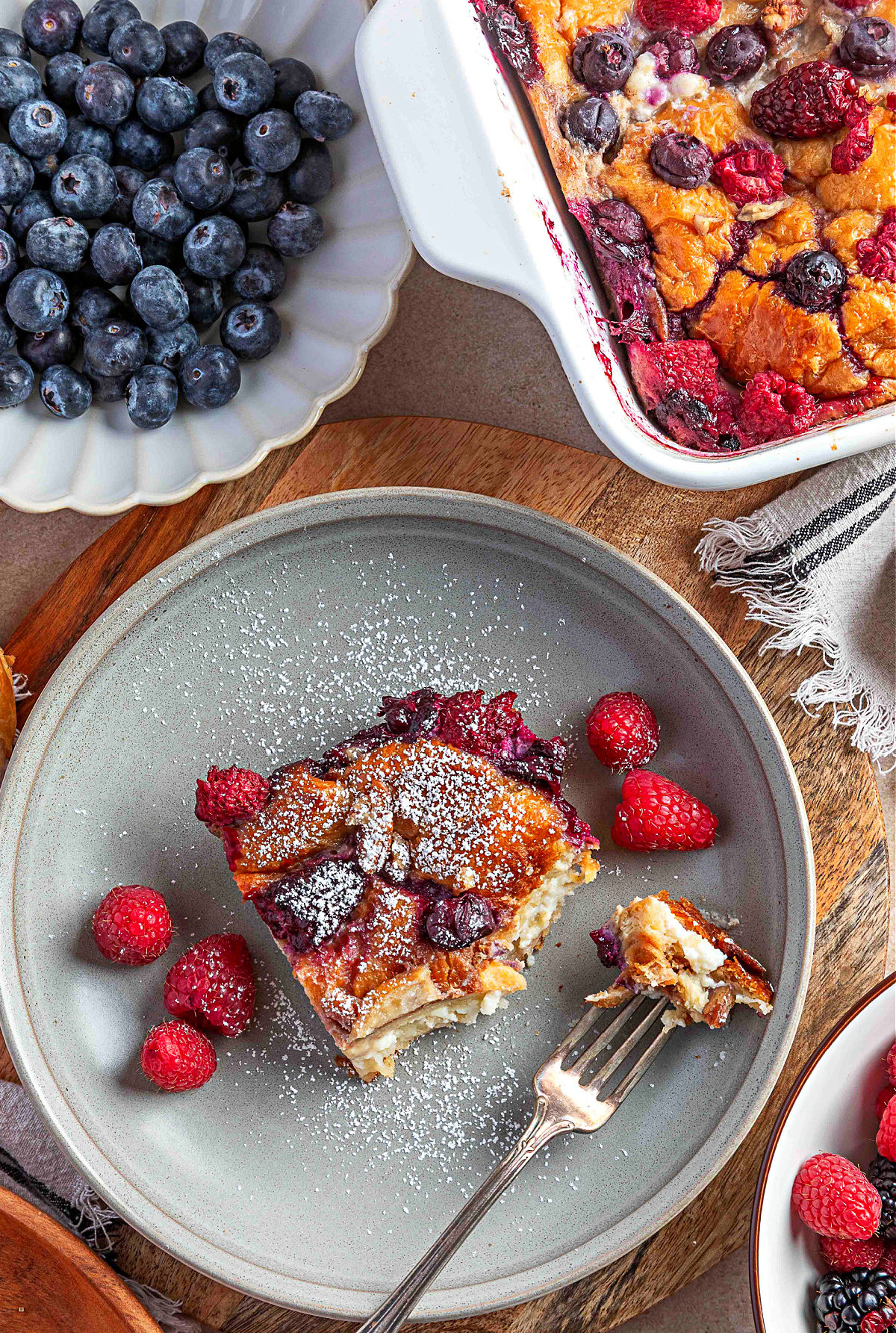 Berry croissant french toast casserole served on a plate with a custardy center, baked berries, and a light dusting of confectioners&rsquo; sugar.