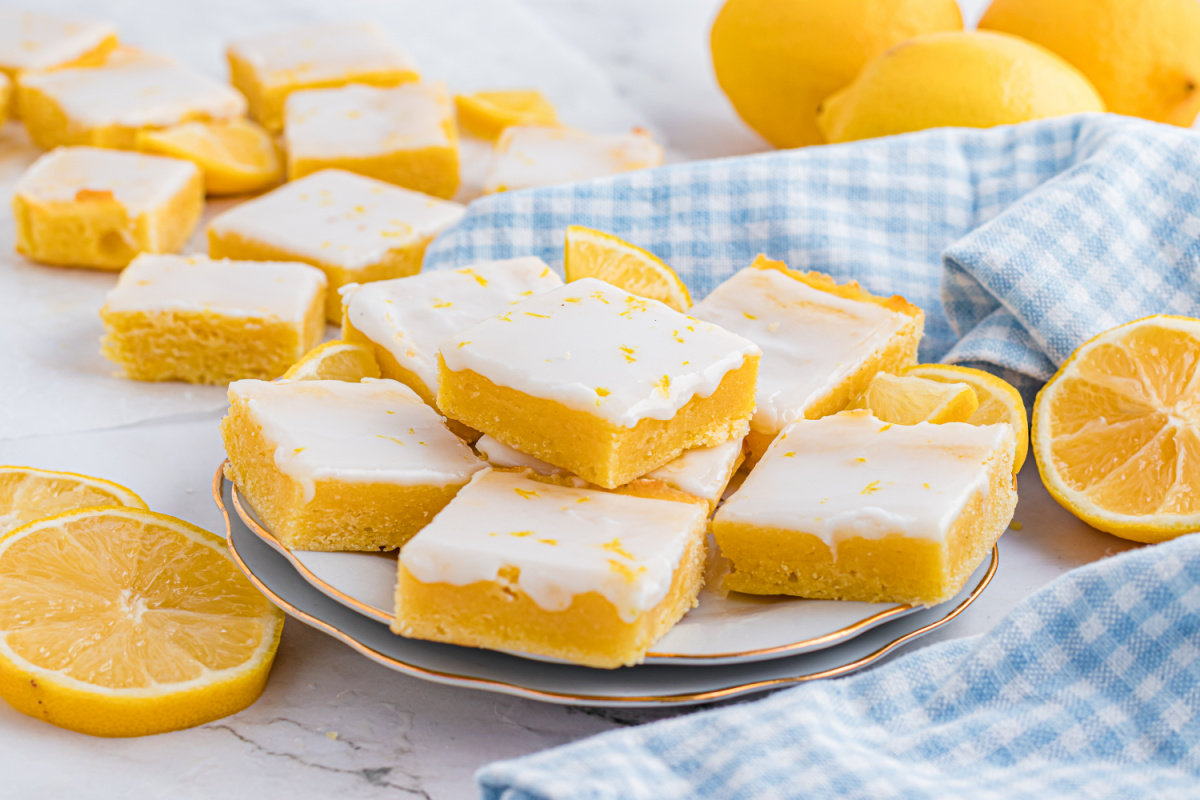 Close-up of glazed citrus dessert bars stacked on a plate, showing a dense lemon crumb and smooth white glaze.