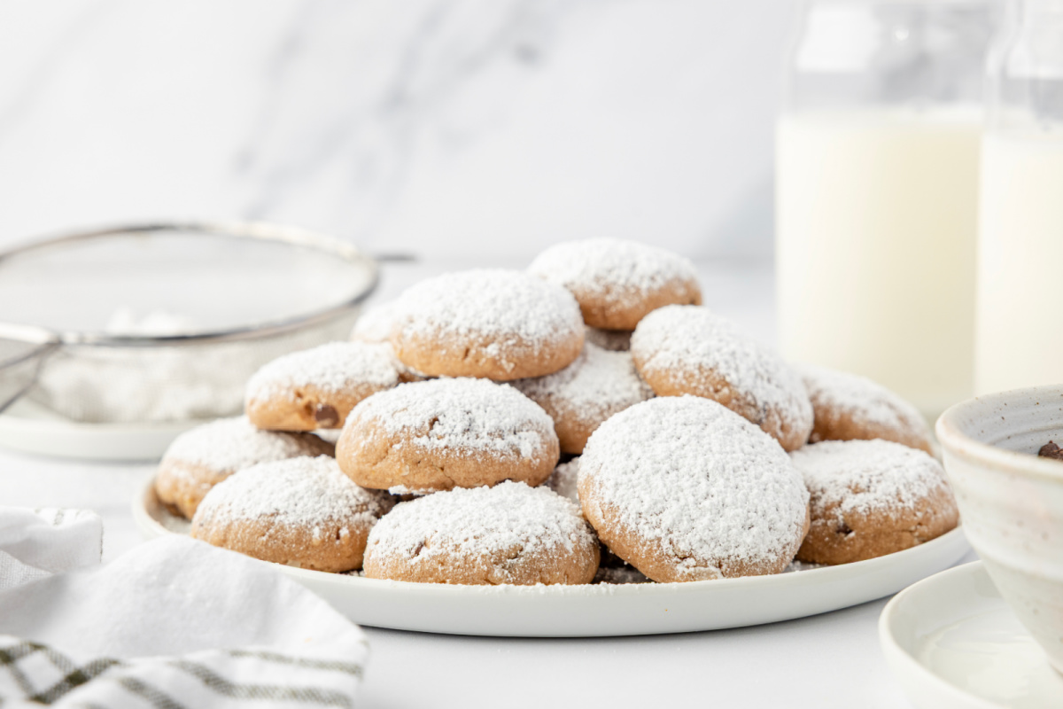 A plate of Danish wedding cookies next to a sugar sifter and bottles of milk on a bright countertop. 