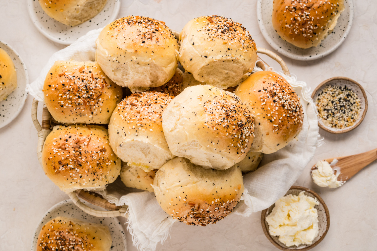 A basket filled with freshly baked rolls topped with sesame seeds, poppy seeds, garlic, and onion, surrounded by small plates of cream cheese and extra seasoning.