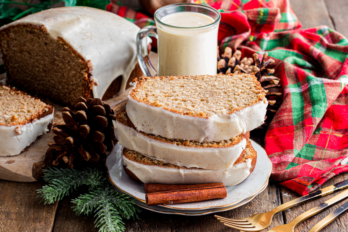 Sliced eggnog bread stacked on a plate with a set glaze, surrounded by pinecones, cinnamon sticks, and a mug of eggnog on a holiday table.