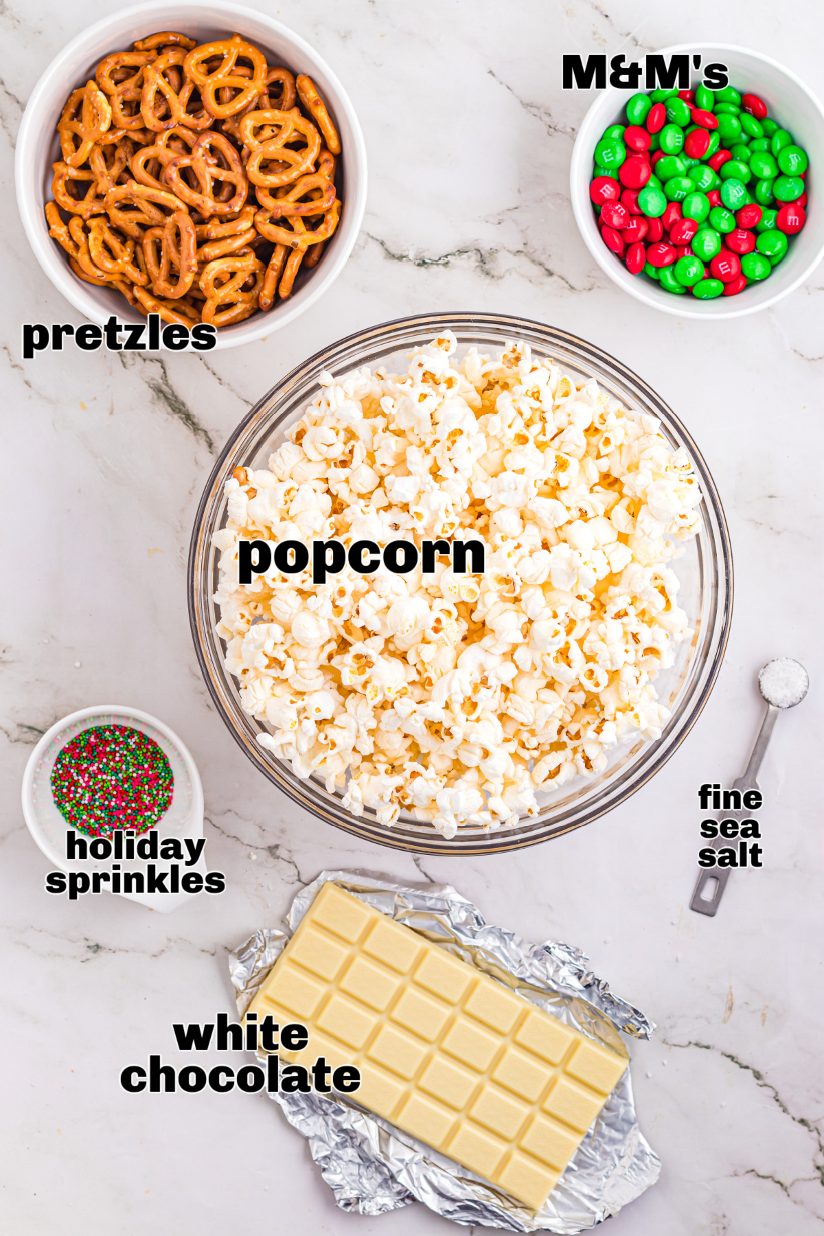 Ingredients for Christmas popcorn mix including plain popcorn, mini pretzels, white chocolate, holiday sprinkles, and candy-coated chocolates.