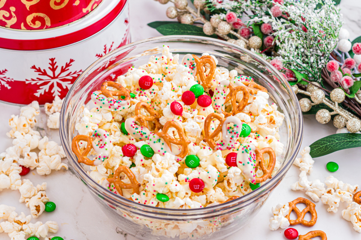 Holiday popcorn mix with white chocolate&ndash;dipped pretzels, red and green candy-coated chocolates, and sprinkles in a glass bowl.