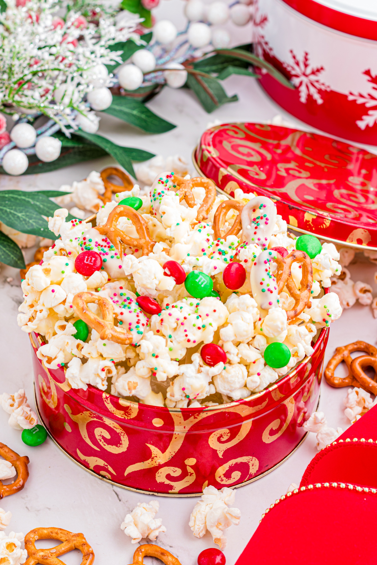 White chocolate popcorn with pretzels, holiday M&M&rsquo;s, and sprinkles piled into a red Christmas tin