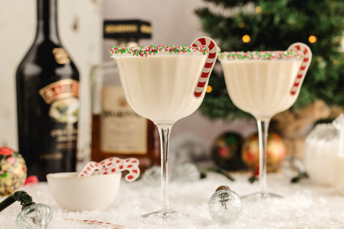 Two creamy holiday martinis with sprinkle rims and candy cane stirrers set on a snowy tabletop, with liqueur bottles and a Christmas tree softly blurred in the background.