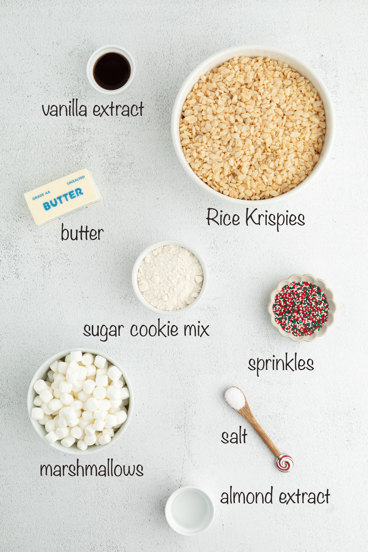 Ingredients for sugar cookie&ndash;flavored Rice Krispie treats arranged on a light surface, including marshmallows, cereal, butter, sugar cookie mix, extracts, salt, and holiday sprinkles.
