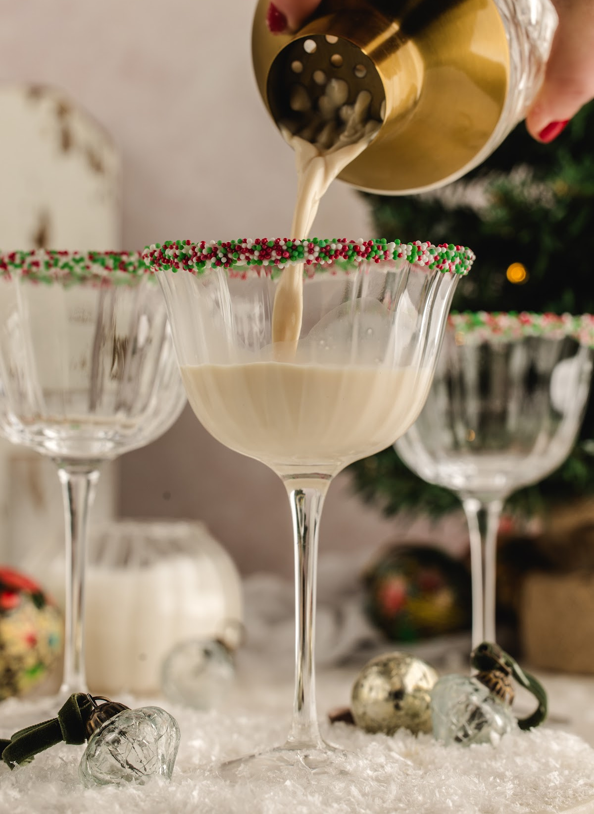 A creamy holiday cocktail being strained from a gold shaker into a sprinkle-rimmed coupe glass, surrounded by ornaments and snowy decorations.