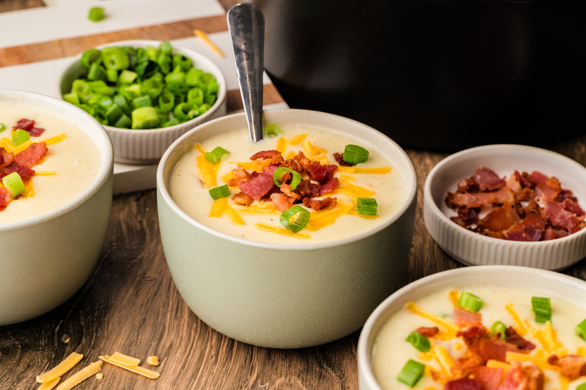 A bowl of bacon cheddar potato soup garnished with scallions, set on a plate beside the pot and ladle.