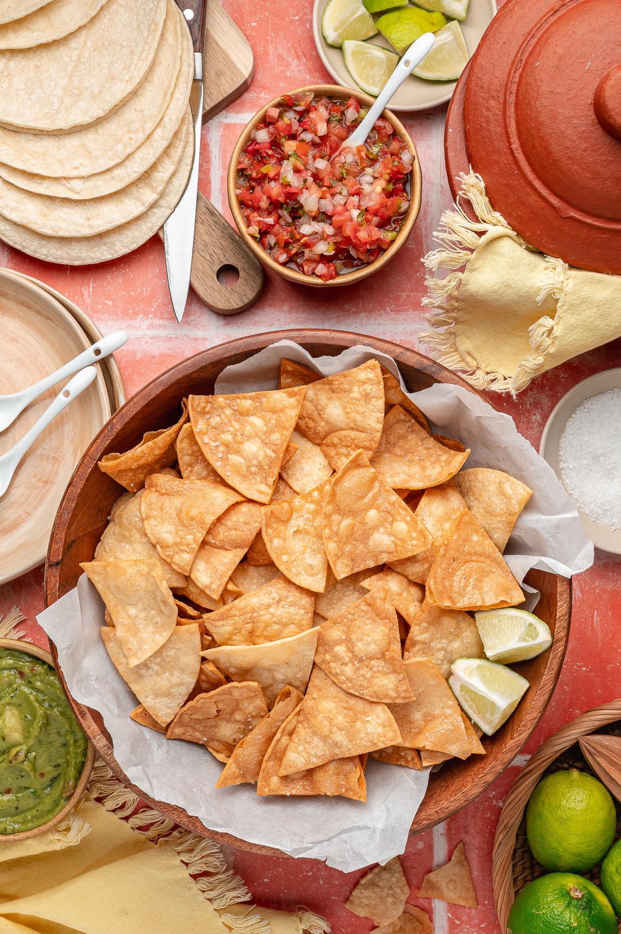 A large wooden bowl of easy fried tortilla chips piled high with blistered, golden wedges, surrounded by fresh salsa, guacamole, and lime.