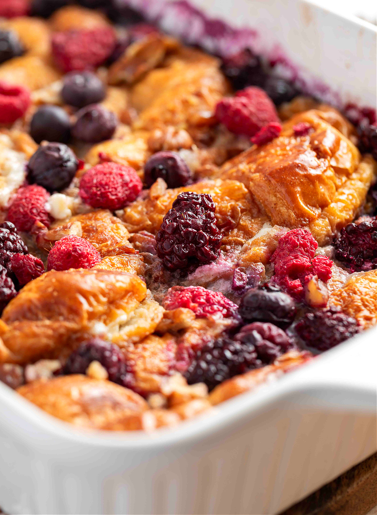 A step-by-step collage showing the process of mixing custard, sweetening cream cheese, stuffing croissants, soaking them in custard, and assembling the French toast casserole with berries and pecans.